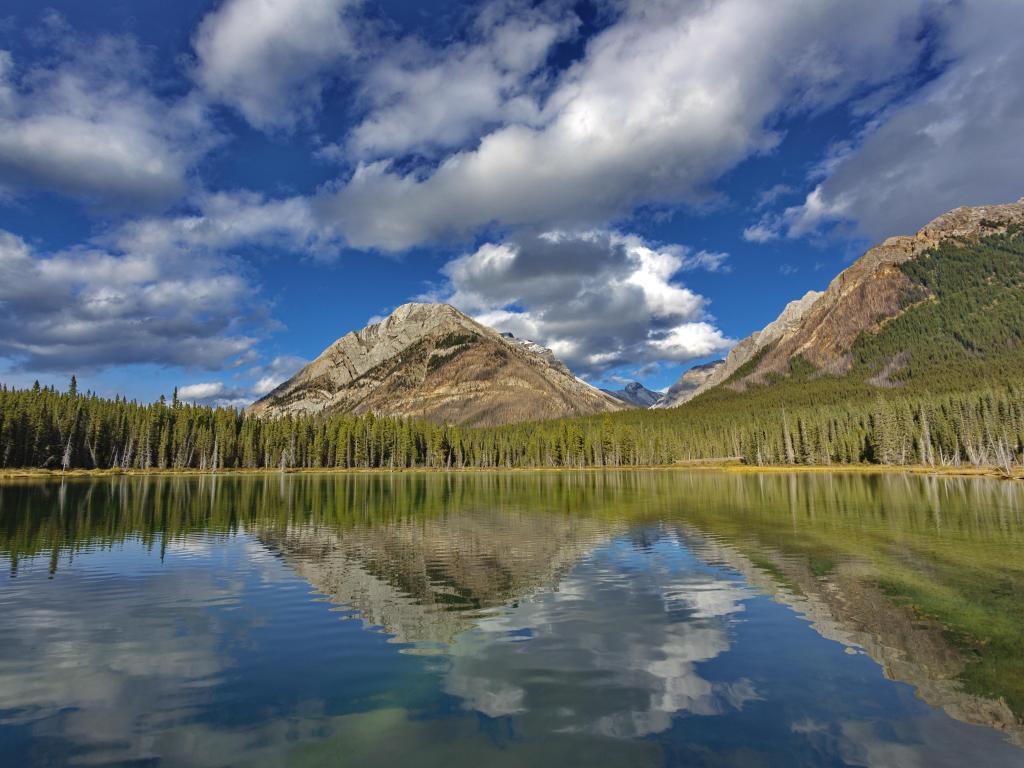 Big blue sky, mountains, and beautiful clouds reflect in still waters of Buller Pond in Spray Valley Provincial Park in Alberta, Canada. 