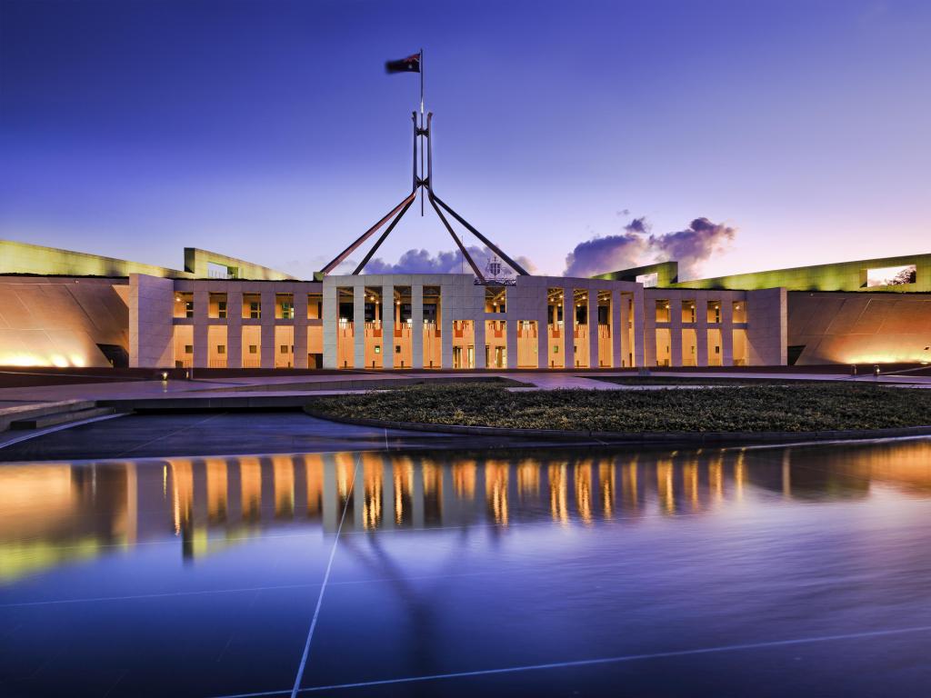 Facade of Australian National Parliament House, Canberra, brightly illuminated and reflecting in blurred water of fountain pond under waving national flag on flagpole at sunset.
