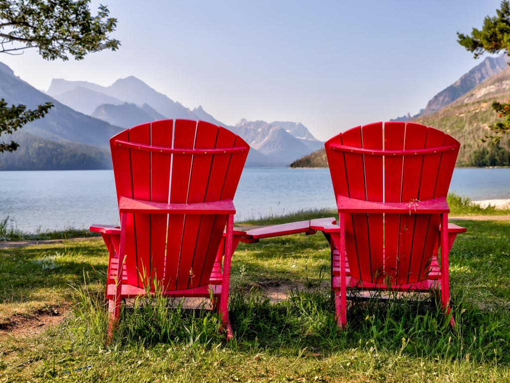 Red chair look over Waterton Lake Marina Point in autumn foliage season sunny morning. Blue sky with colourful clouds reflect on the lake water surface. 