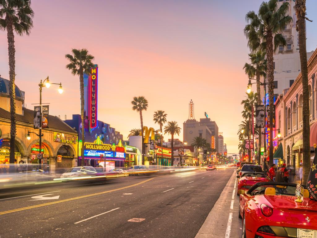 Los Angeles, California, USA with traffic and sports cars on Hollywood Boulevard at dusk with palm trees and iconic buildings in the distance.