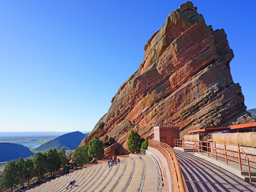 A picturesque view of the Historic open-air Red Rocks Amphitheatre with a couple of people sitting in the benches enjoying the view of the mountains from afar early in the morning in Denver