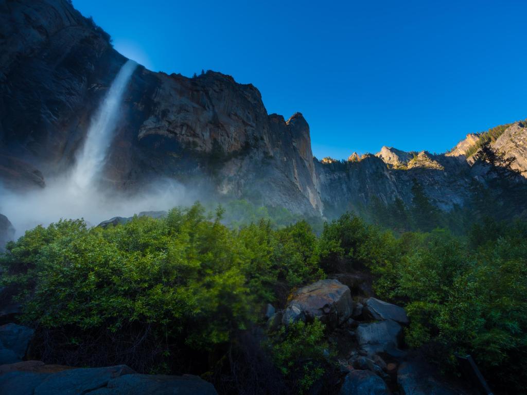 Crashing waters falling from Bridalveil Falls surrounded by lush green forest and blue skies