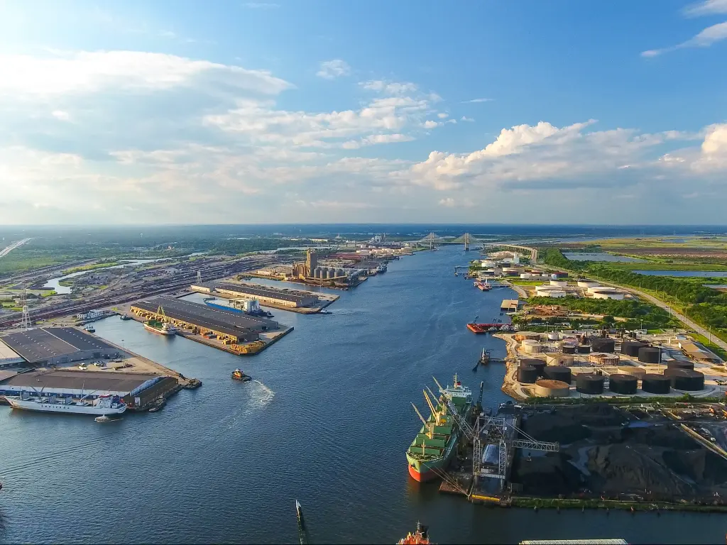 An aerial view of Mobile river and the port of Mobile, Alabama with ships dock in the harbor, green trees, and sunny weather with a sheet of white and gray clouds spread in the blue sky