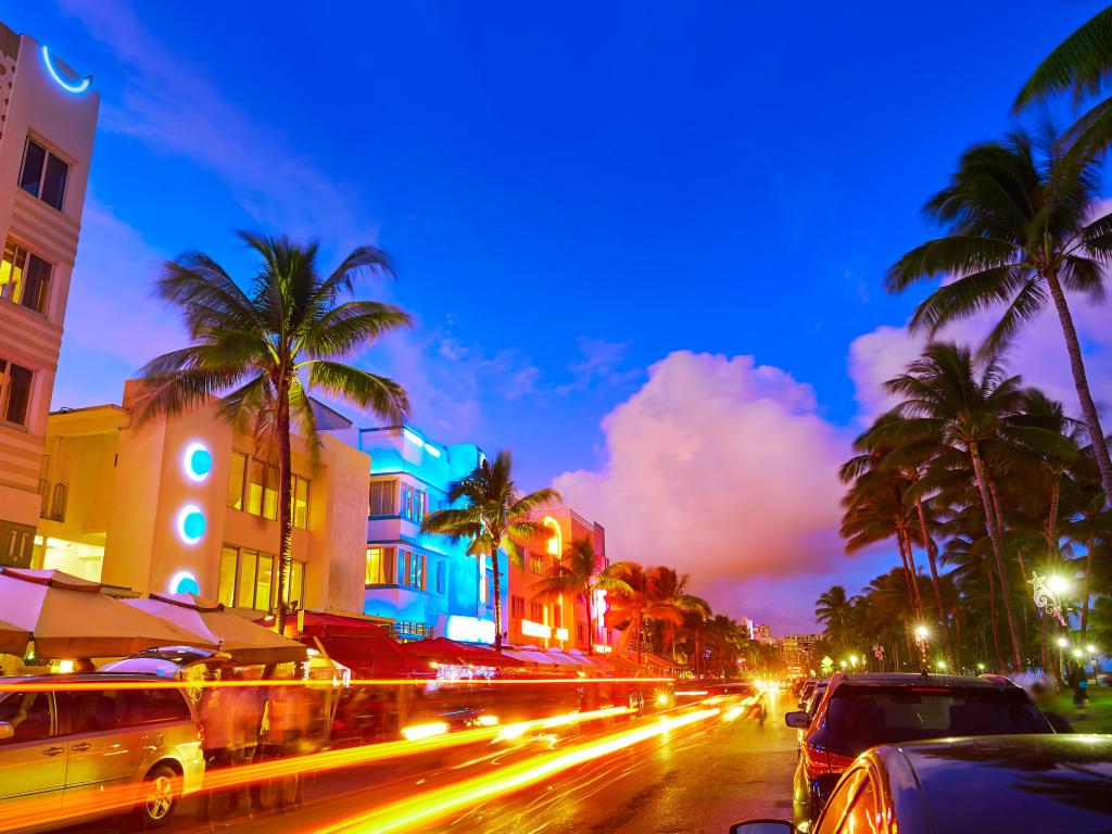 Art Deco buildings illuminated in neon colors with car lights and palm trees