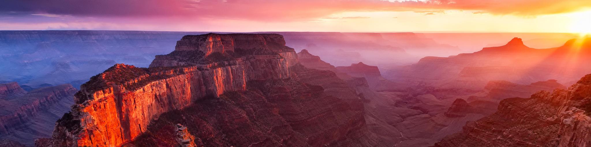 The Grand Canyon, USA taken at sunset with a dramatic sky off reds and yellows casting shadows on the epic rock formations in the foreground and the valley below.