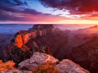 The Grand Canyon, USA taken at sunset with a dramatic sky off reds and yellows casting shadows on the epic rock formations in the foreground and the valley below.