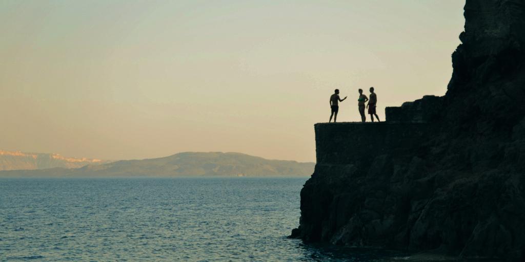 Three people stand on a cliff in Amoudi Bay, Santorini, getting ready to jump into the water