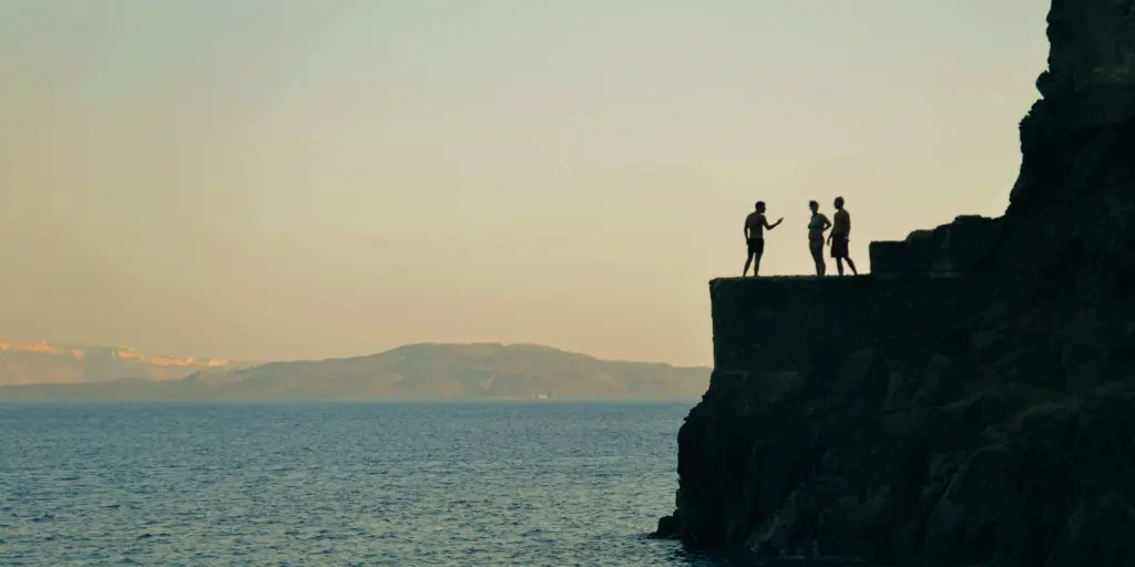 Three people stand on a cliff in Amoudi Bay, Santorini, getting ready to jump into the water