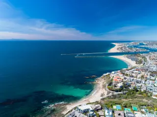 Aerial drone shot over Newport Beach in Orange County, with coastal homes, blue skies, beaches and harbor entrance in view.
