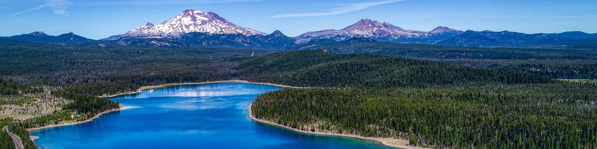 Aerial panorama of Elk Lake near Bend, Oregon, with the deep blue and turquoise lake sitting in front of snow capped mountains, under a blue sky
