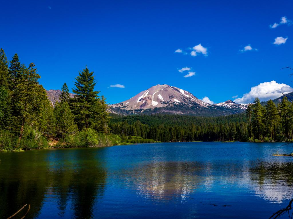 Lassen Volcanic National Park, California with Lassen Peak in the distance reflected in Manzanita Lake in the foreground and surrounded by tall trees under a blue sky.