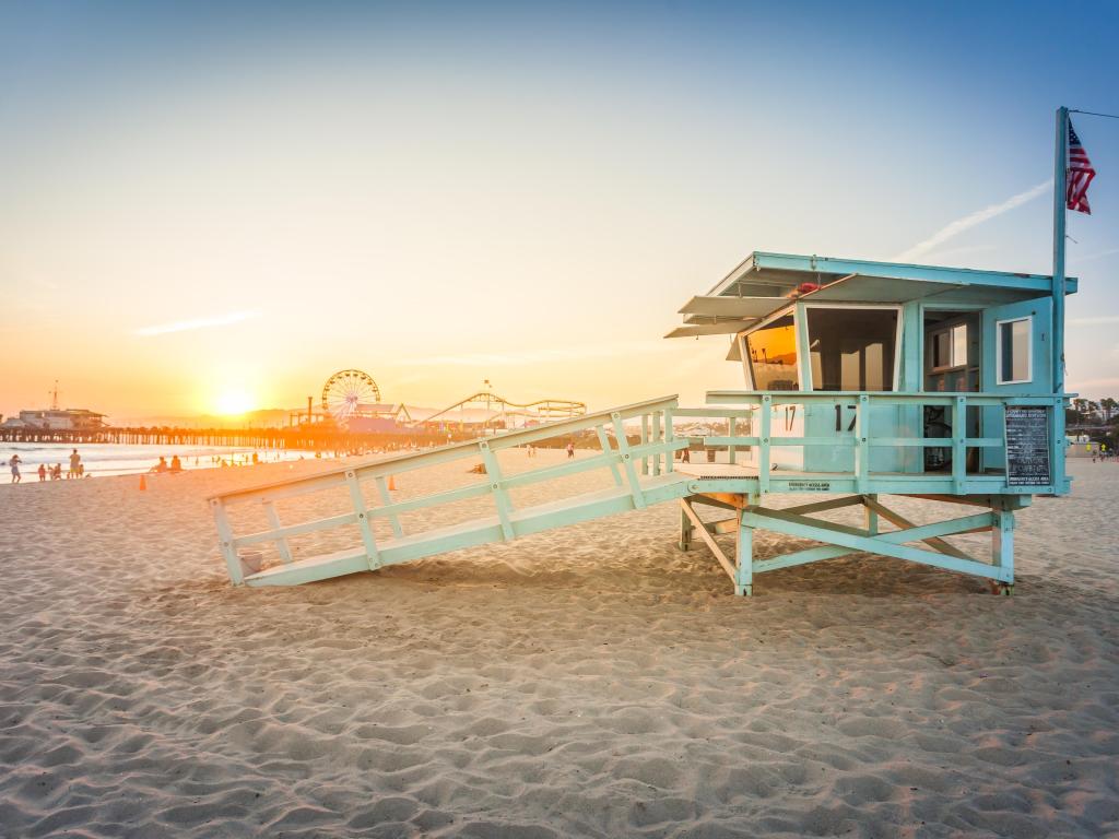 Sandy beach with sun setting on the horizon, with vibrant blue lifeguard cabin in the foreground and amusement park in the background