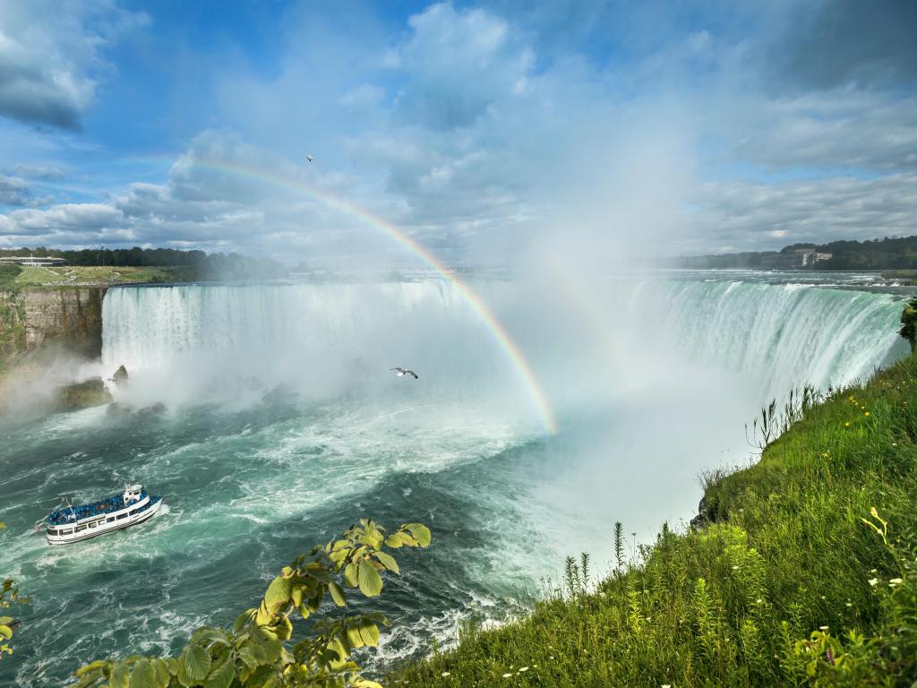 Rainbow shining in the spray rising from the Horseshoe Falls at Niagara Falls on a sunny day, with a boat passing by