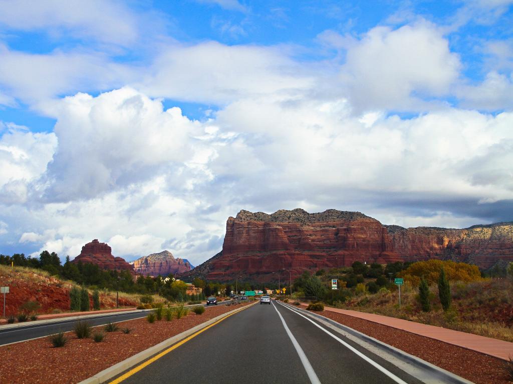 An image of a road that is part of SR 179 also known as Red Rock Scenic Byway in Sedona, Arizona that has mountains on the sides in red color during a sunny day.