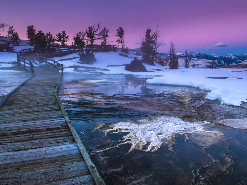 Path leading past partly frozen water with snowy mountains in the distance and a pink sunset