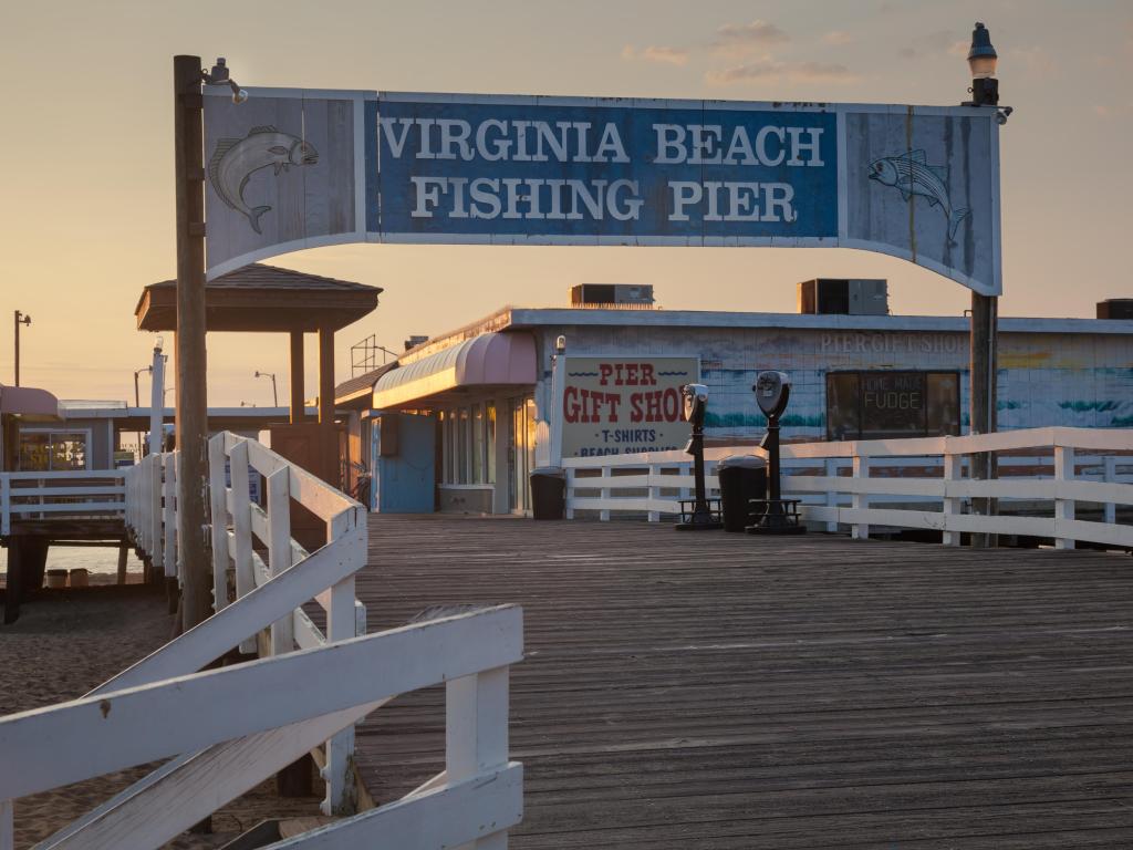Signposted archway entrance to Virginia Beach Fishing Pier at dusk, with a gift shop sign in the background