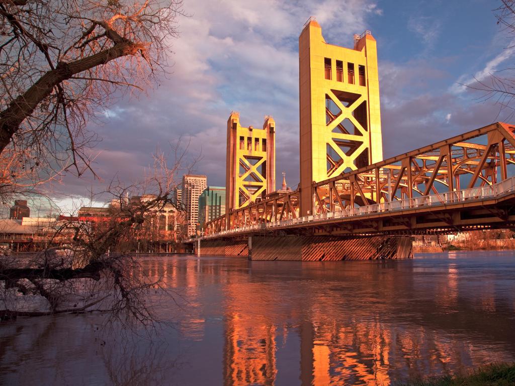 Sacramento, California with the old bridge on one side and a tree hanging over the rive in the foreground at dusk.