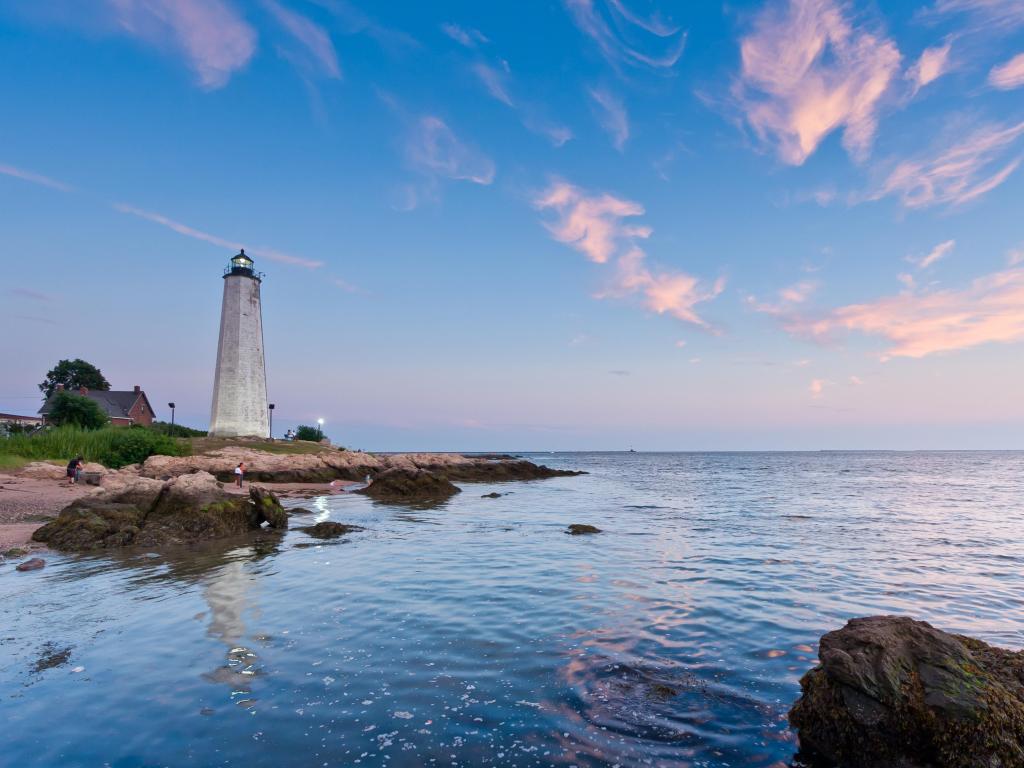 New Haven, Connecticut with the lighthouse at dusk, a beach surrounding the lighthouse and calm water in the foreground. 