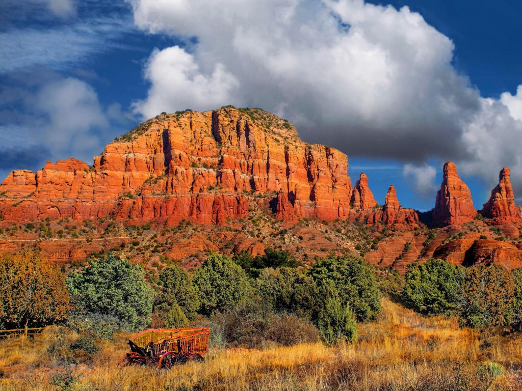 Coconino National Forest, Sedona, Arizona, USA with a red mountain range landscape in the background against a cloudy blue sky, trees and a cart in the foreground.