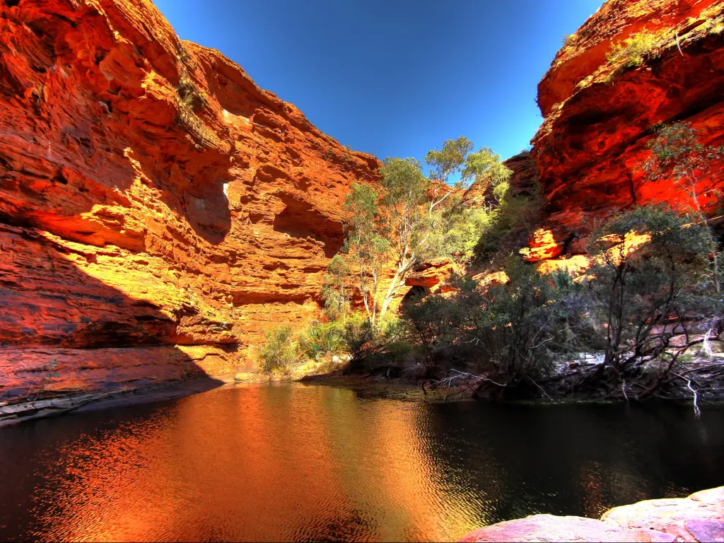 Sun shining between red rocks onto water running through Kings Canyon, with blue sky above