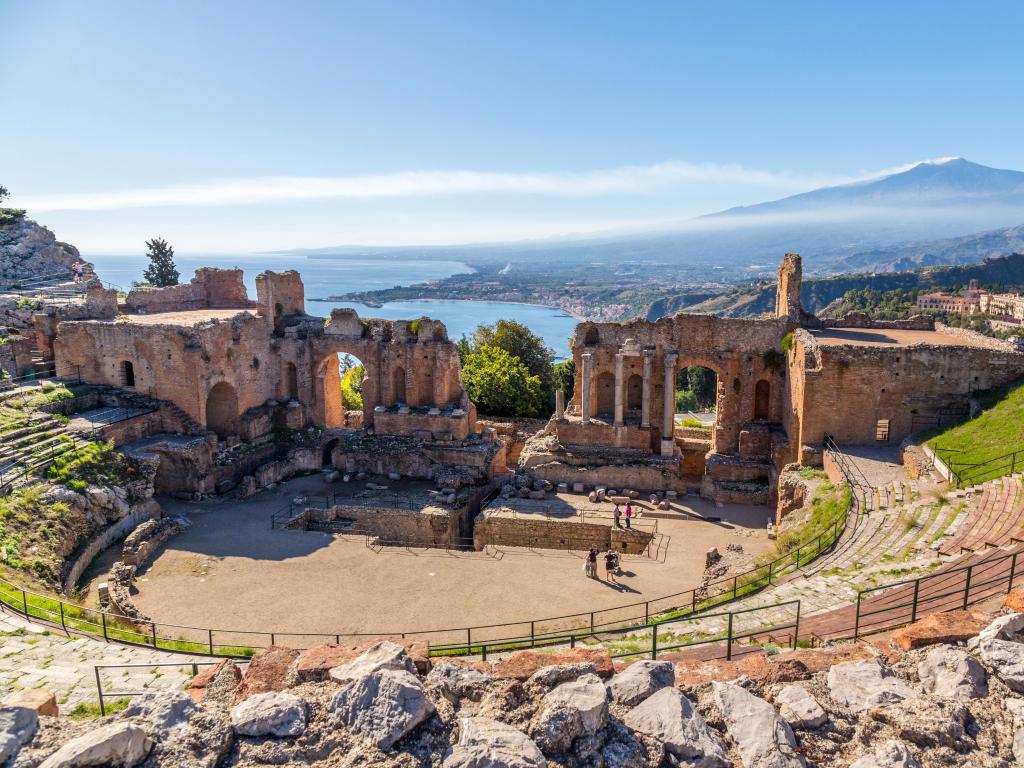 Ruins of the Ancient Greek Theater in Taormina, Sicily with the double smoke tail of the Etna extending over the the Giardini-Naxos bay of the Ionian Sea in the morning sun shine.