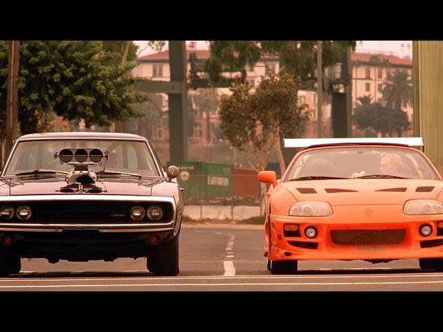 Two cars, one black and one orange, with the main characters of the Fast and Furious movie, Dom and Brian, about to start a race.
