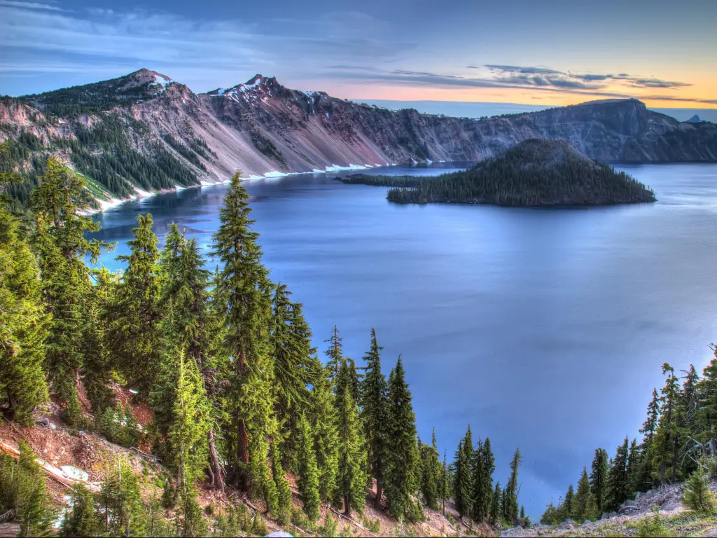 Crater Lake National Park, Oregon with tall trees in the foreground and a vast lake leading to mountains in the background at sunset. 