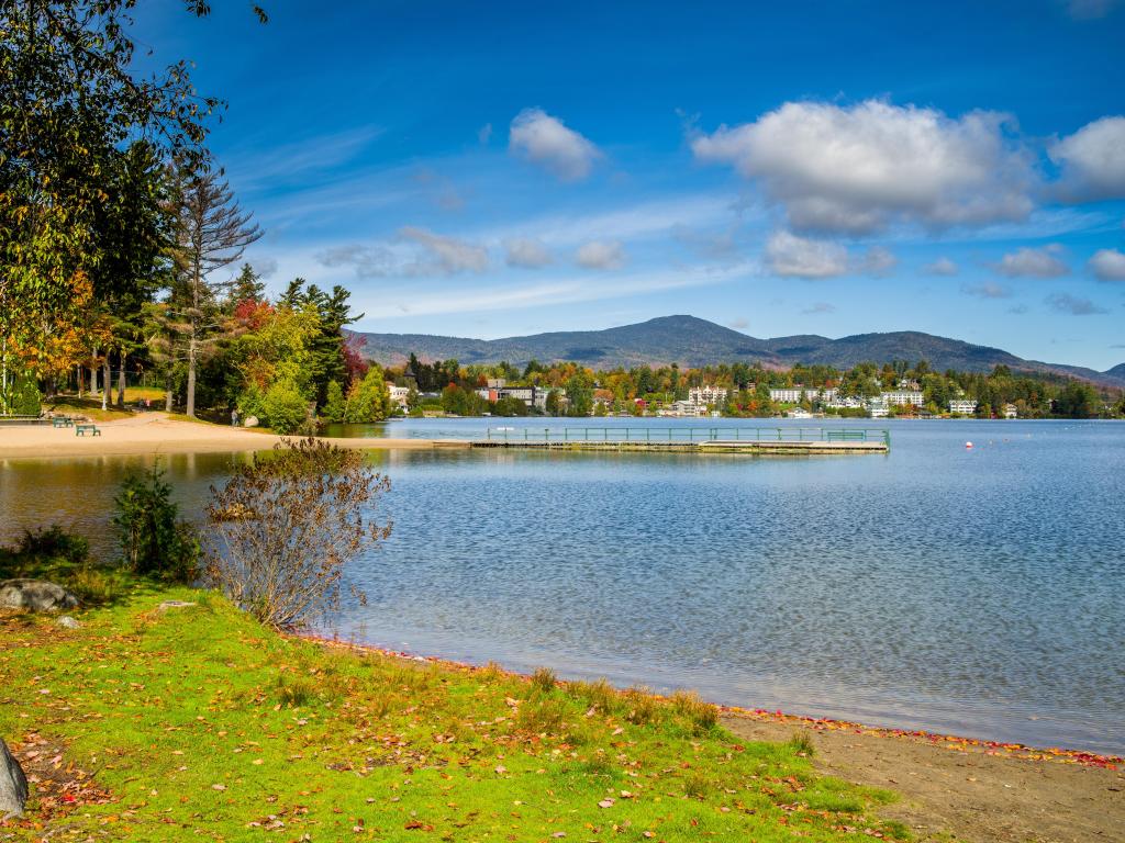 View across Mirror Lake, with blue skies and mountain backdrop, and lush green parkland surrounding the shoreline, Upstate New York