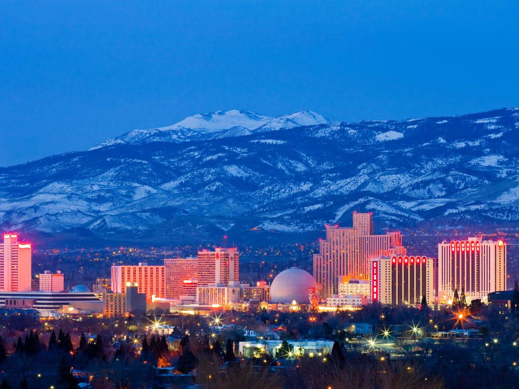 Brightly lit highrise buildings of downtown Reno with snow-covered mountains rising up in the background