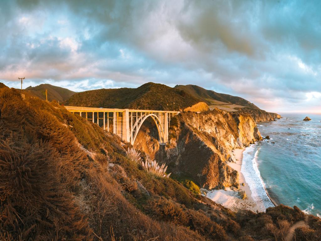 Panoramic view of Bixby Creek Bridge crossing high cliffs with blue sea and golden sunset light
