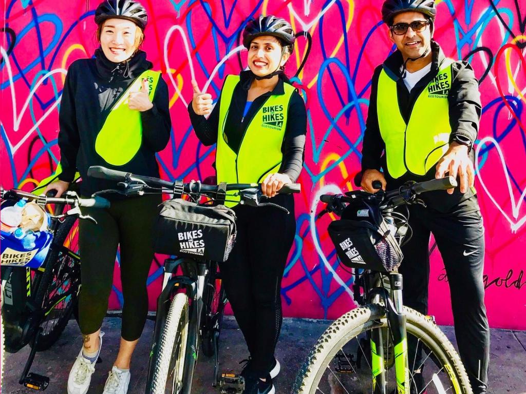 Colorfully dressed bikers holding their bikes and giving the camera thumbs-up with a bright pink background