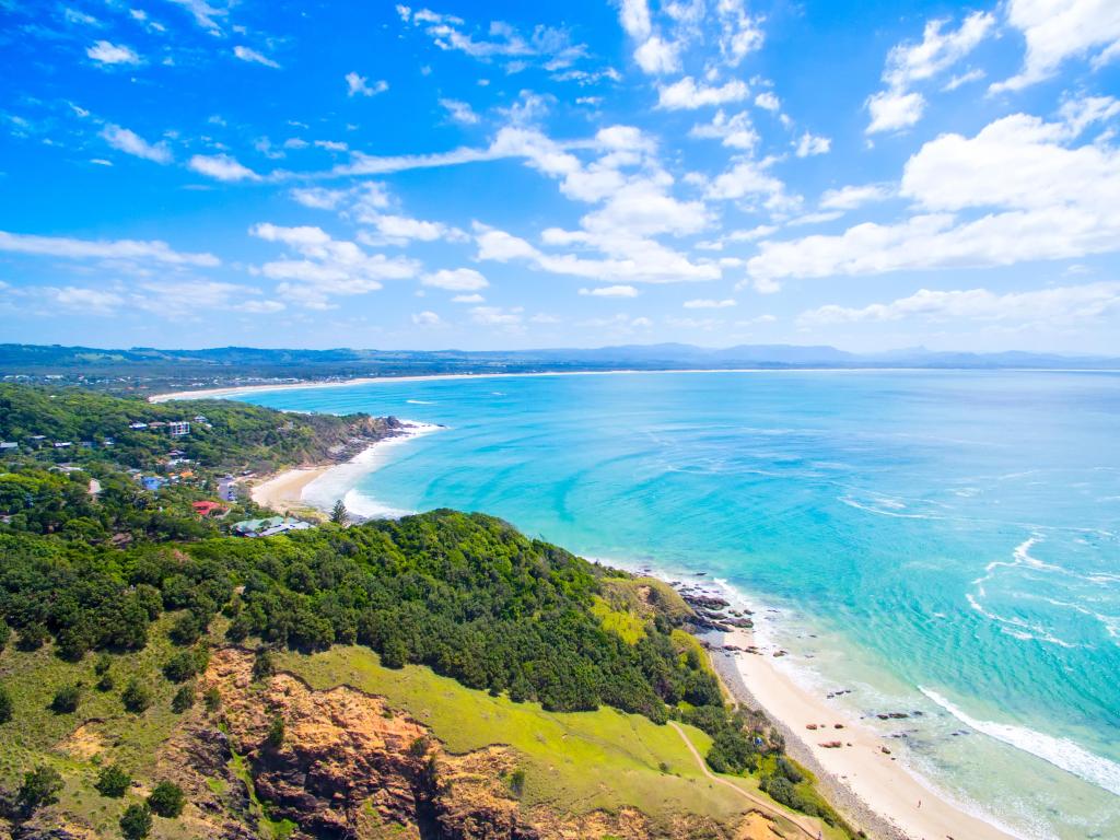 Byron Bay, Australia coastline taken as an aerial shot showing the calm clear waters encased by beaches and tree-topped hills.