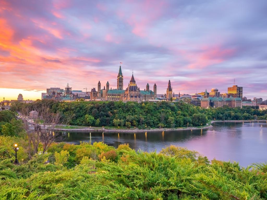 Parliament Hill in Ottawa, Ontario, Canada taken at sunset with trees in the foreground and calm water. 