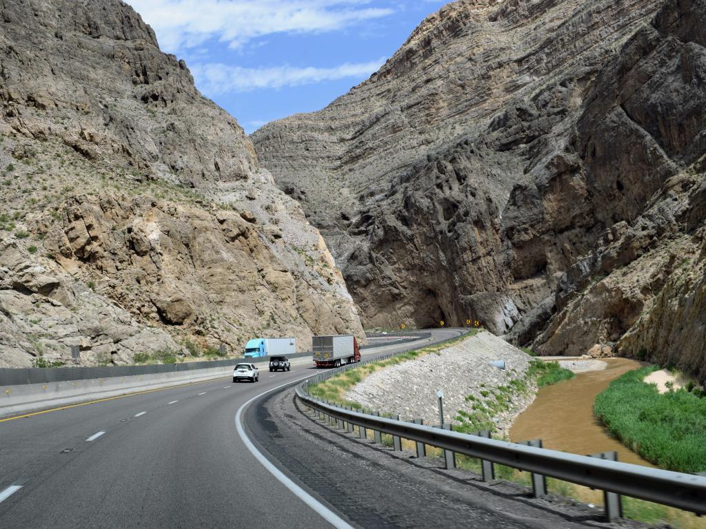 A winding road of I-15 that is running alongside Virgin River with the mountains ahead on both sides with some cars travelling.