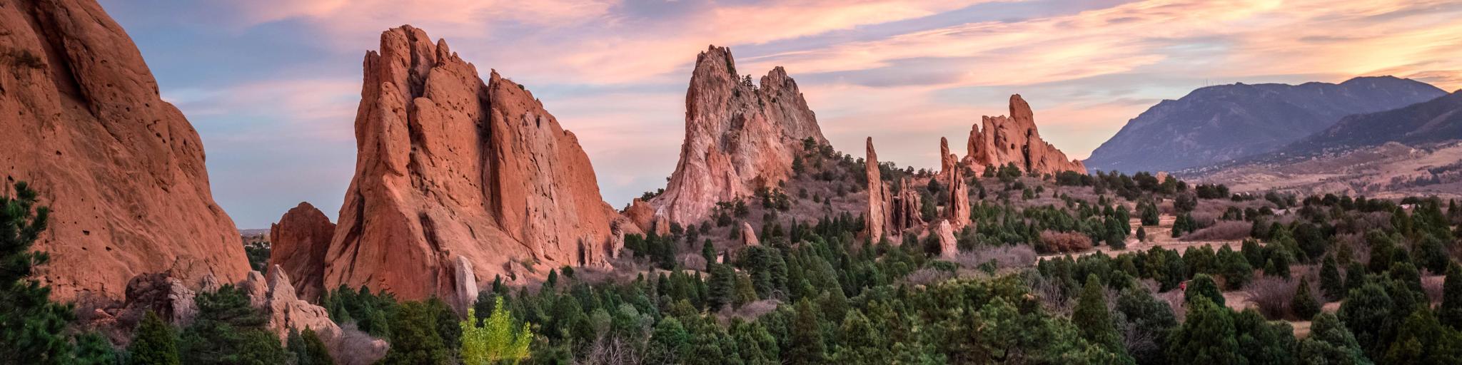 Garden of the Gods, Colorado Springs at sunset with green trees in the foreground and the jagged rock formations in the background.