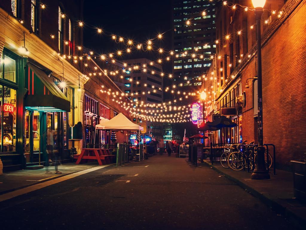 Tables for street food laid out on a narrow street with colourful shop fronts and lights strung across the alleyway