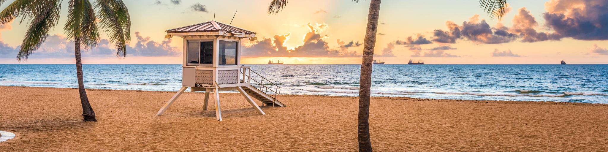 Fort Lauderdale, Florida, USA with the beach in the foreground and a few palm trees, a wooden platform in the middle and the sea in the background at sunset. 