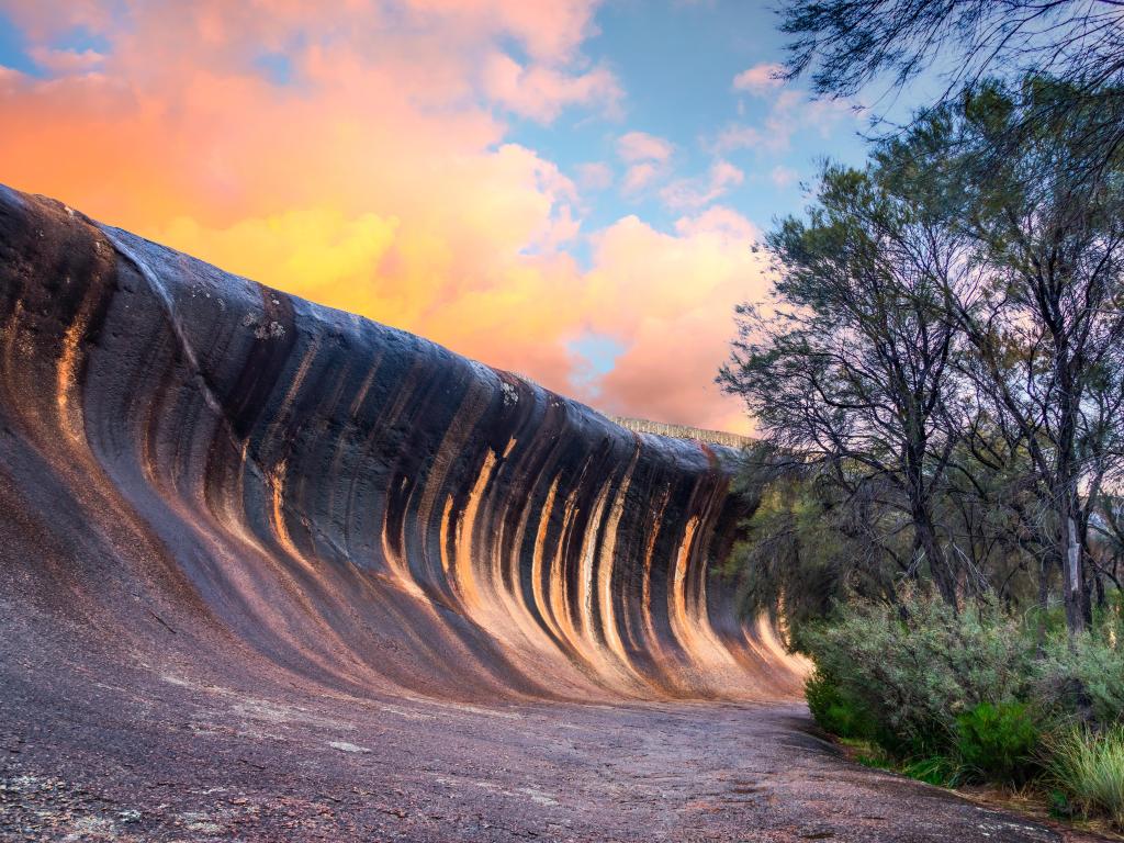 Spectacular wave-shaped rock formation crosses the image, with a vivid orange and pink sunset sky
