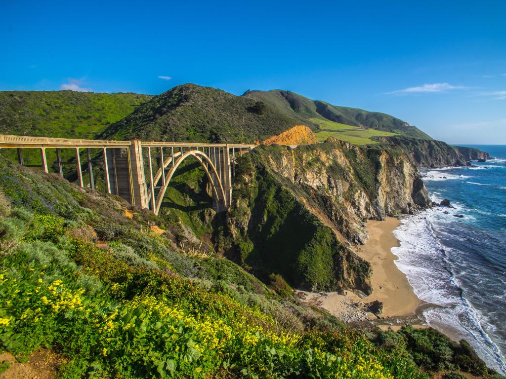 Big Sur, California, USA with a view of Bixby Bridge surrounded by green cliffs over the Pacific Ocean on a sunny clear day.