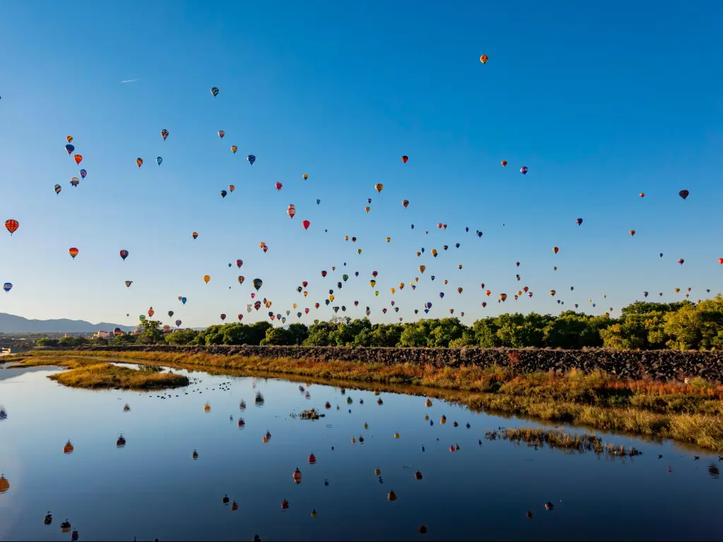 Many brightly coloured hot air balloons fly through blue sky above a lake, reflected in the water below