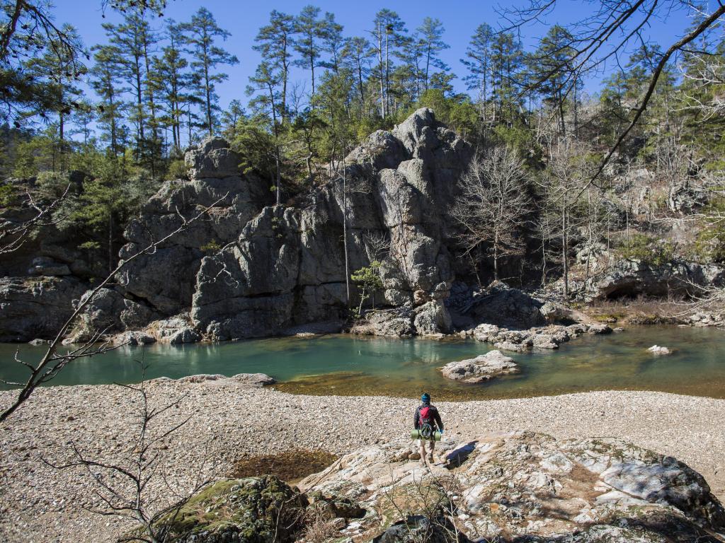 Ouachita Mountains, Arkansas with a hiker walking towards a river with rock formations in the background and trees in the distance. 