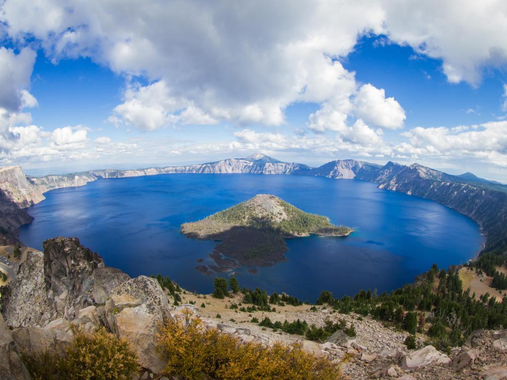 Crater Lake, Oregon taken as a wide angle view of the lake form the top of Watchman's Peak, rocks and trees surrounding on a cloudy blue sky.