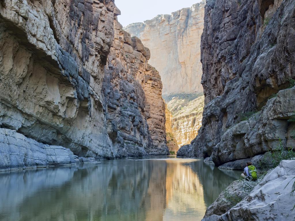 The Rio Grande River running through Santa Elena Canyon at Big Bend National Park in Texas. A hiker rests by the riverbank in the lower right, facing away from the camera.