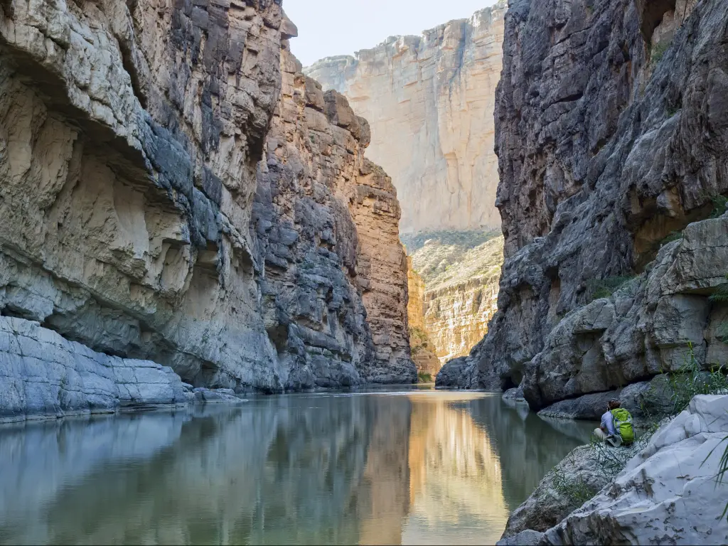 The Rio Grande River running through Santa Elena Canyon at Big Bend National Park in Texas. A hiker rests by the riverbank in the lower right, facing away from the camera.
