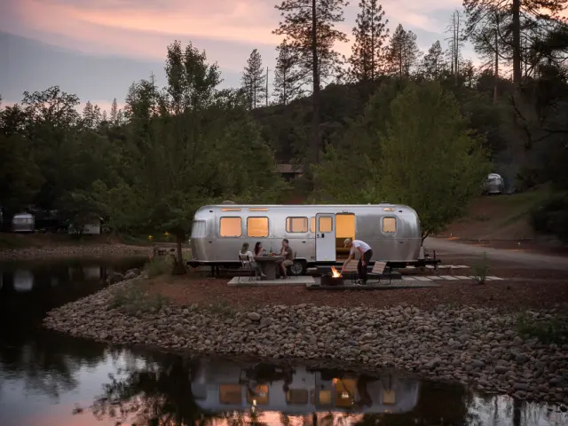 Unique RV Airstream accommodation nestled in the forest, with visitors enjoying the lit firepit as the night draws in