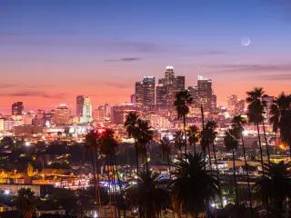 Los Angeles Skyline in the distance at sunset, with palm trees in the foreground, with the moon rising faintly behind