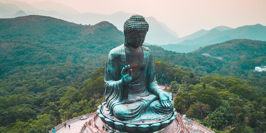 A view of the Tian Tan Buddha, Hong Kong, at sunset with visitors walking around it and the mountains in the background