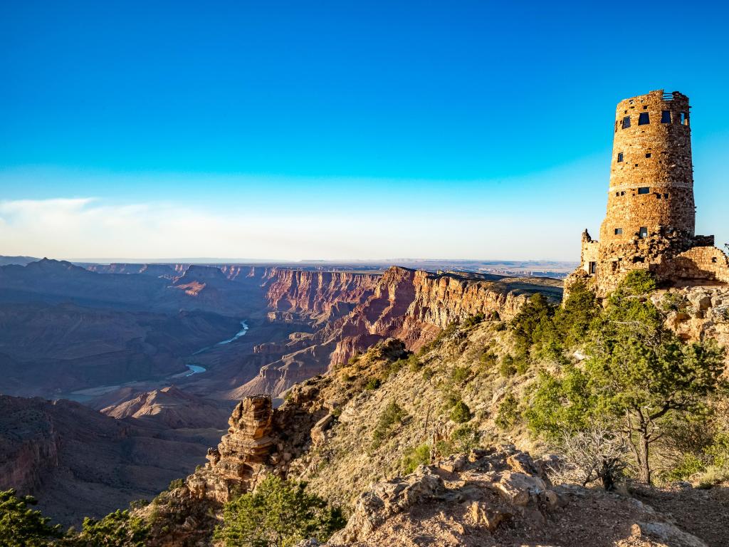 Grand Canyon Village, USA with the Grand Canyon in the distance and an old ruin watchtower in the foreground over a blue sky.