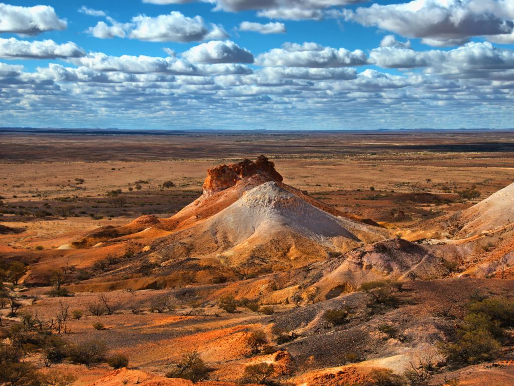 Coober Pedy, Australia with orange rocky terrain in the foreground and flat plains in the distance above a blue cloudy sky. 