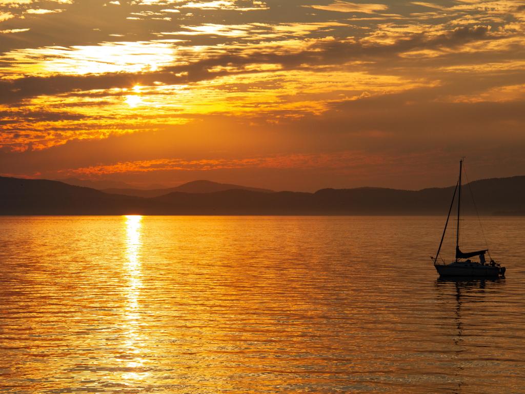 Lake Champlain, Vermont, USA with a sailboat in the foreground, mountains in the distance taken at sunset with yellow and orange hues. 